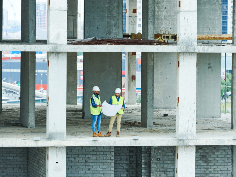 Two men in helmets and waistcoats reading construction scheme while standing on concrete floor of unfinished building